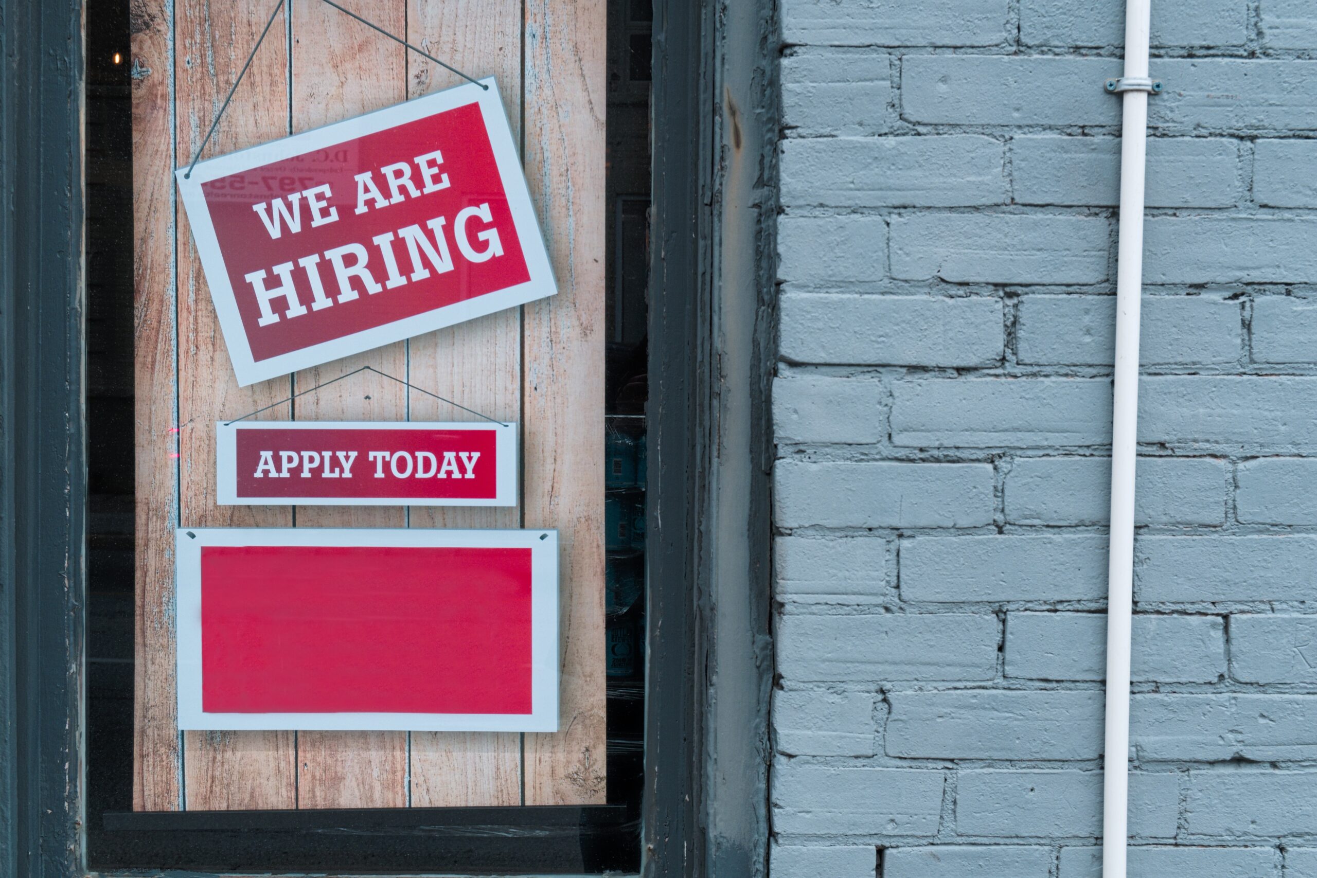 A wooden door in a white-brick painted wall with a red sign saying "we are hiring"