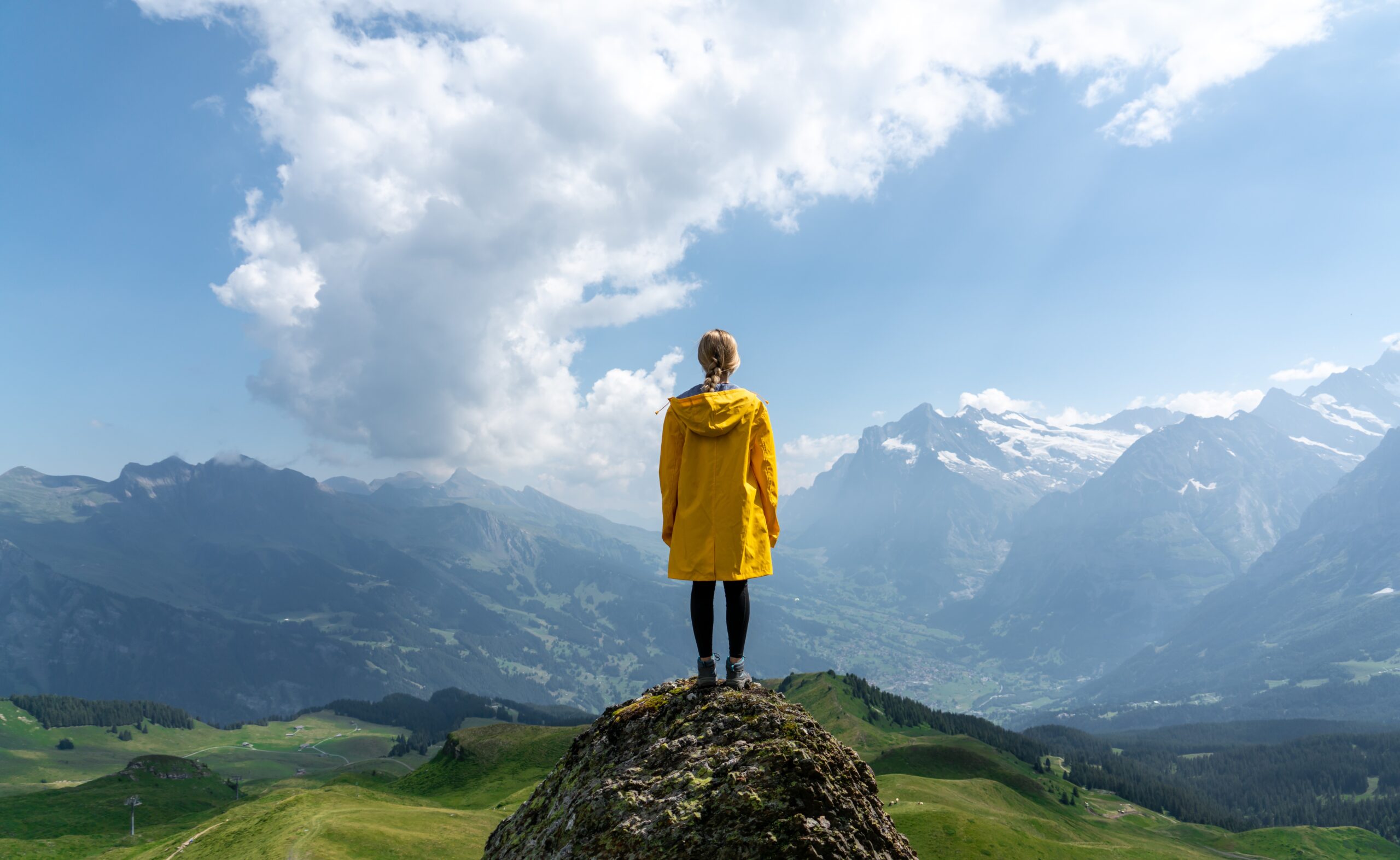 Woman in a yellow jacket standing on a high rock facing away from the camera, and looking over large green fields and snow-capped mountains