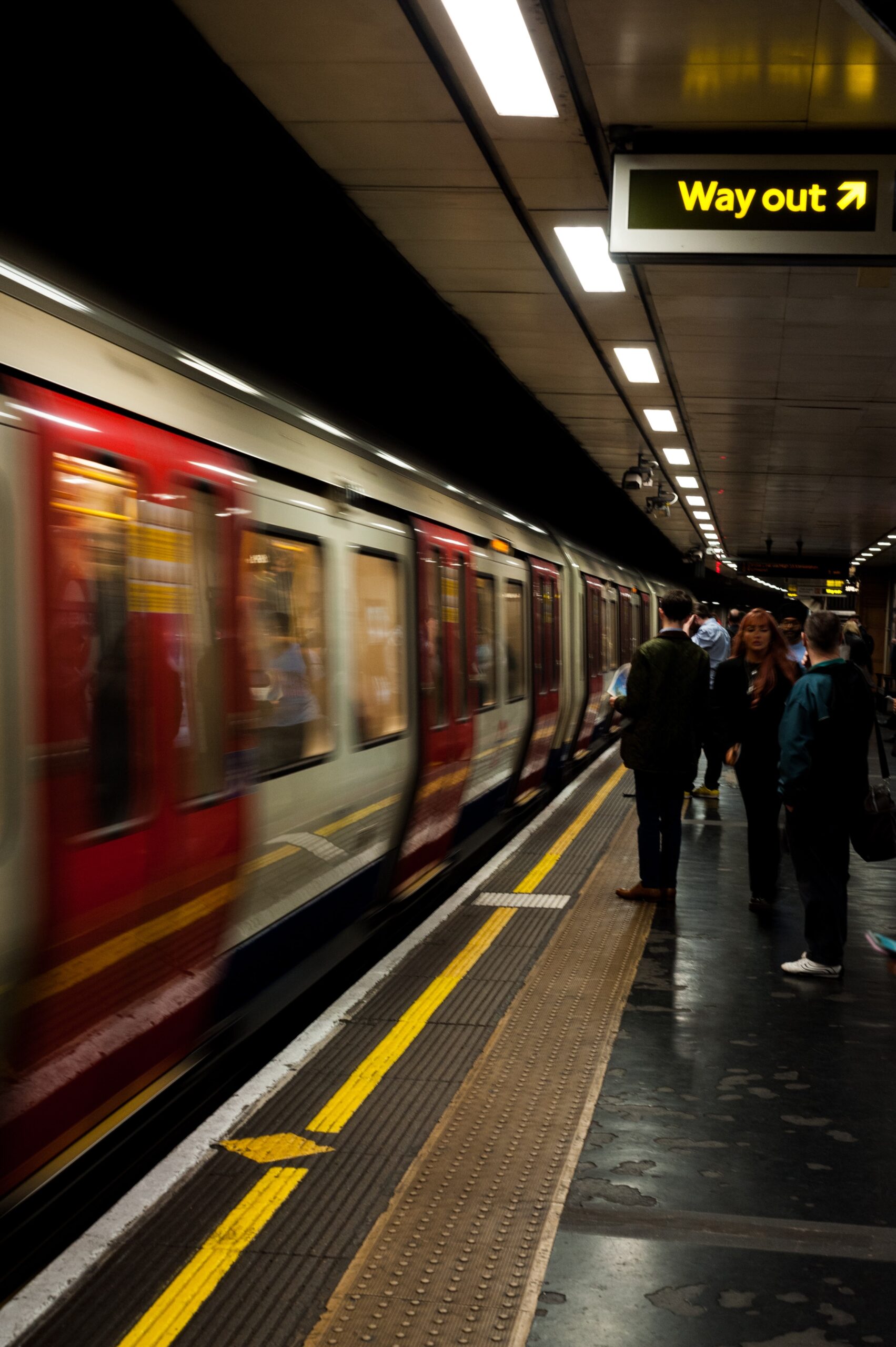 A busy train platform with a train rushing past and a sign overhead reading "way out"