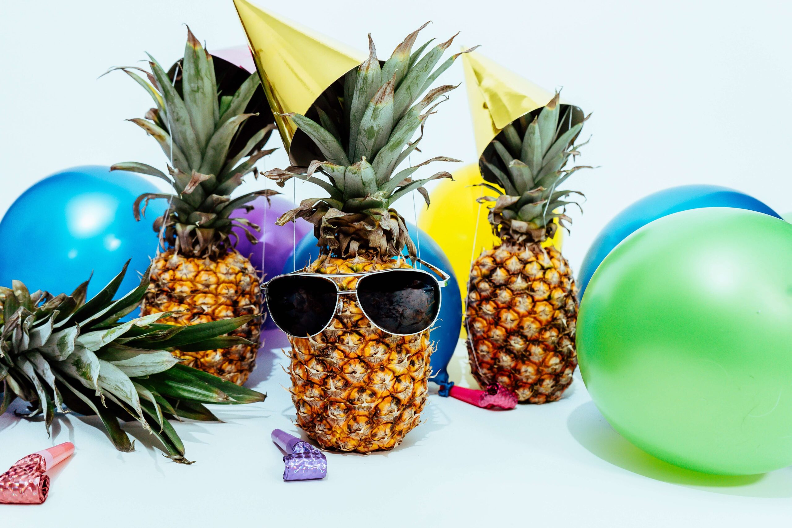 Three pineapples all wearing party hats and one with sunglasses on surrounded by balloons and party poppers