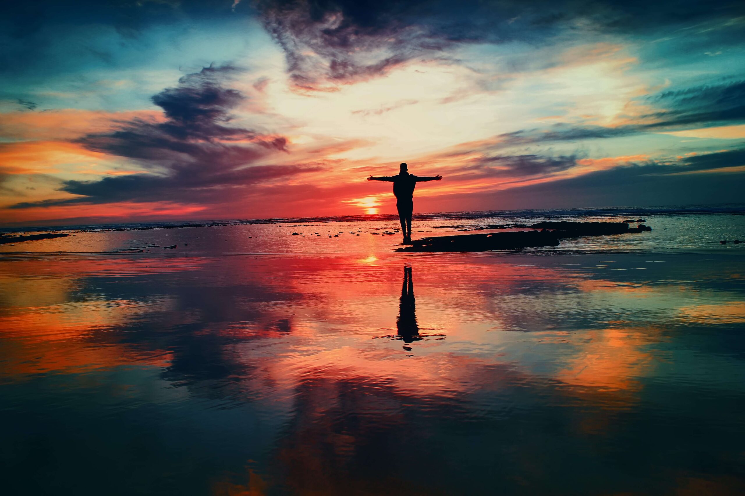 A person stood with their arms outstretched in the distance on a beach at low tide with a stunning sunset of grey, black, grey and oranges lighting up the sky and sea