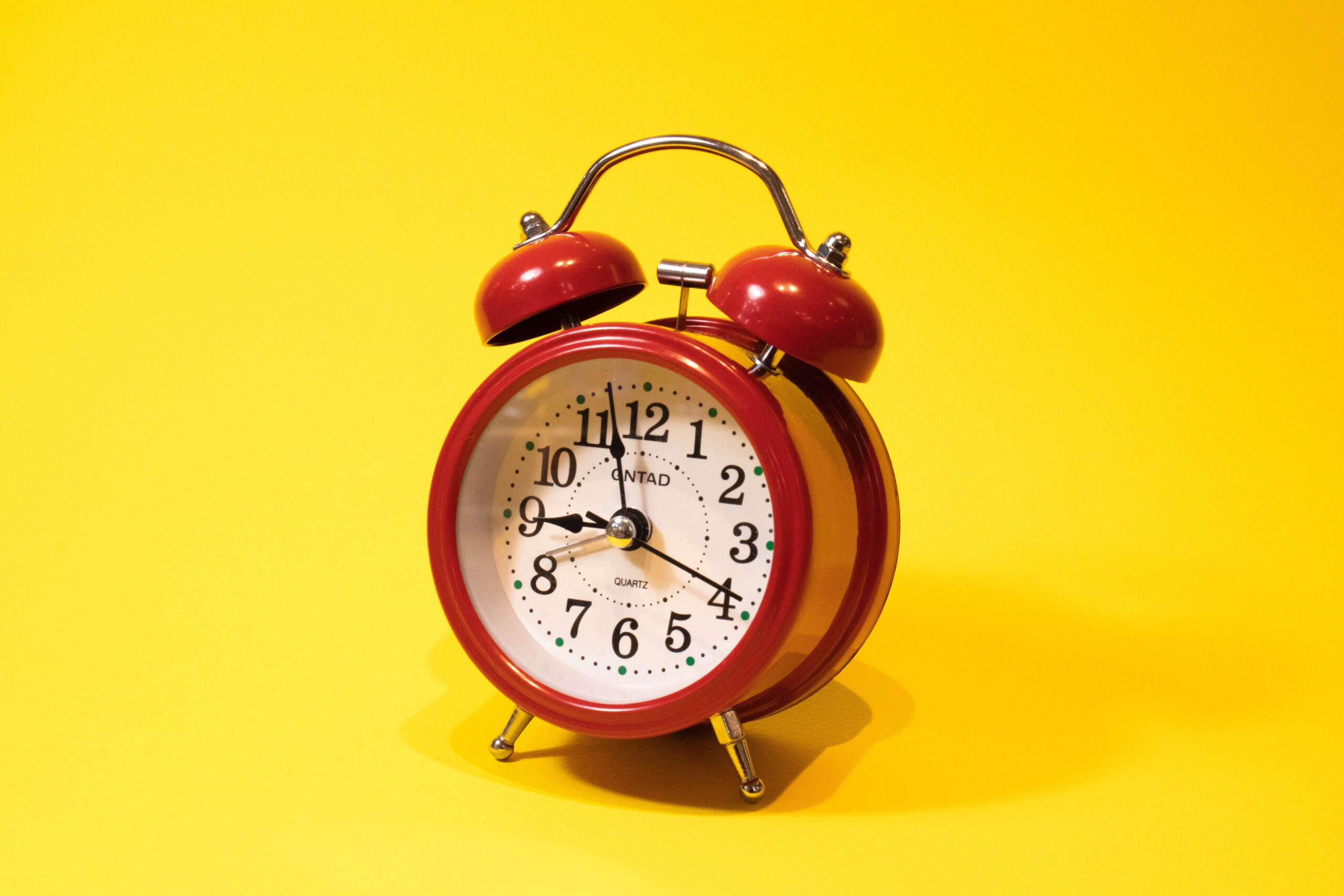 A red clock with a round face and the bells on top set against a vivid yellow background