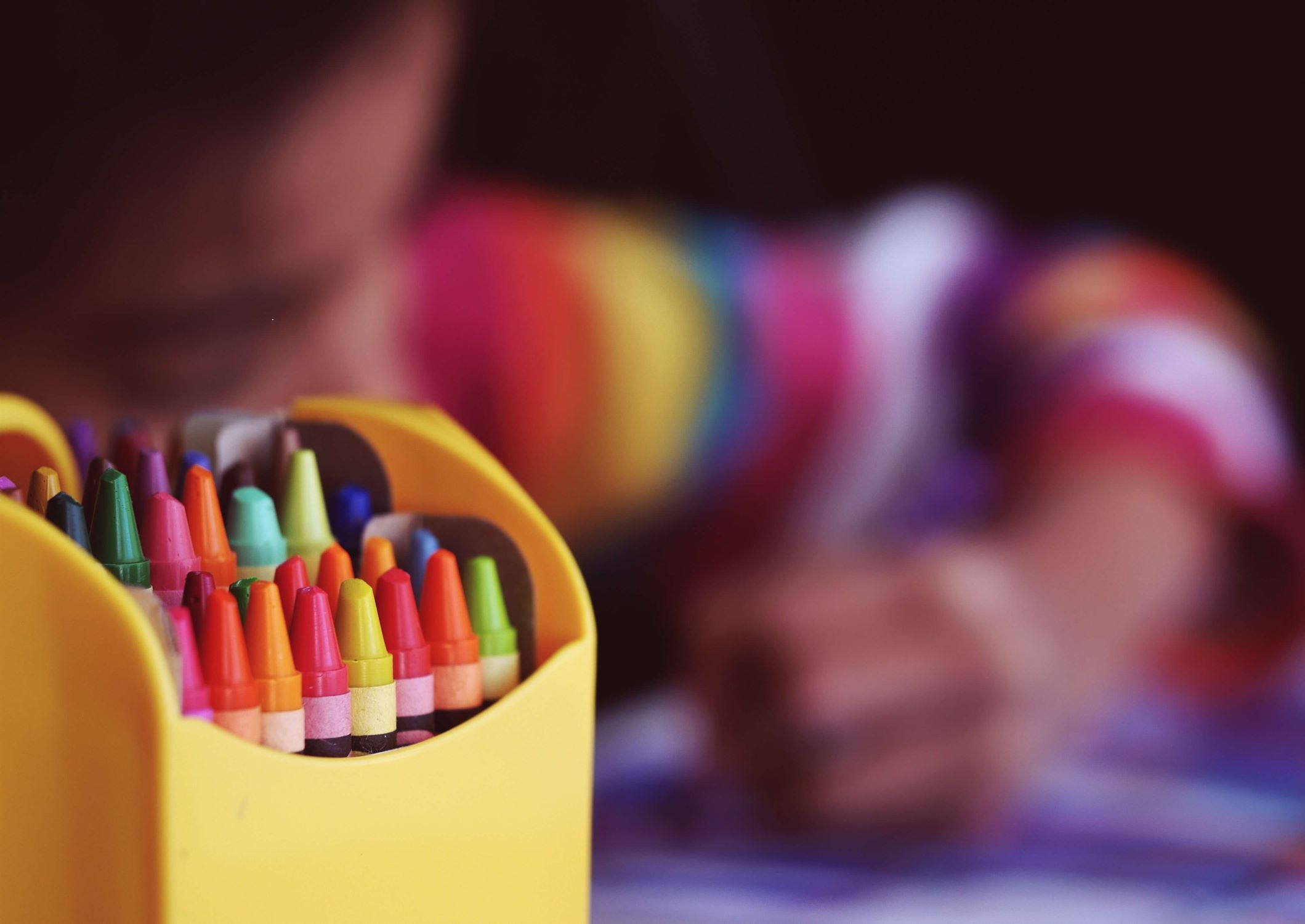 A pack of coloured crayons in the front of the picture with a child colouring blurred in the background