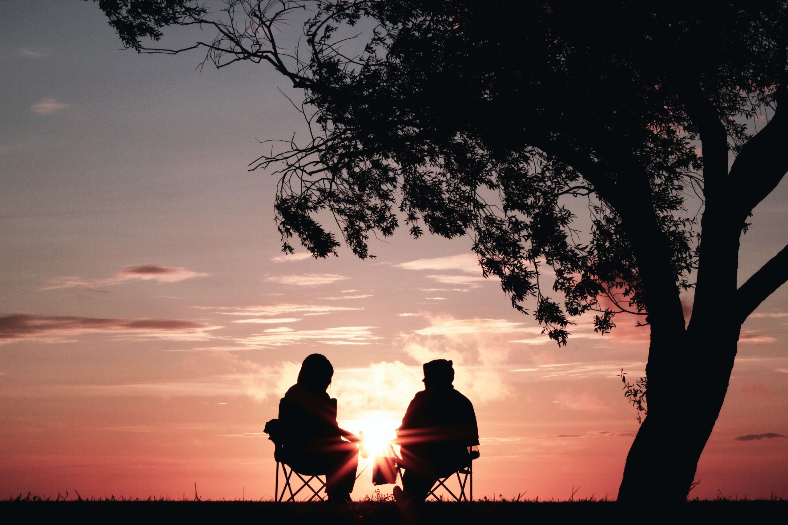 Two people sitting on chairs next to a chair in silhouette looking at the sun setting