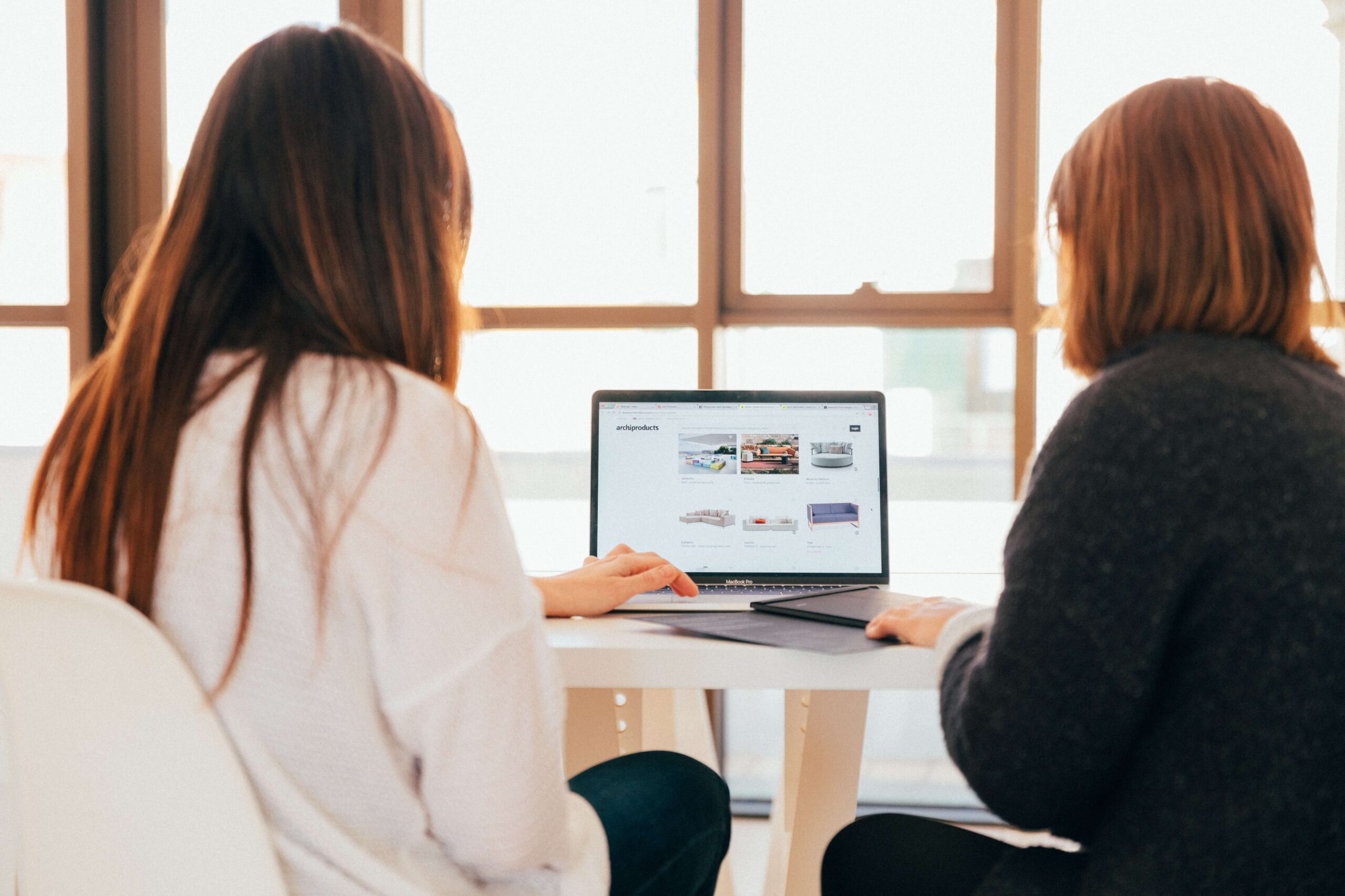 Two women with their backs to the camera, sat down at a table looking at a laptop together in front of a big window