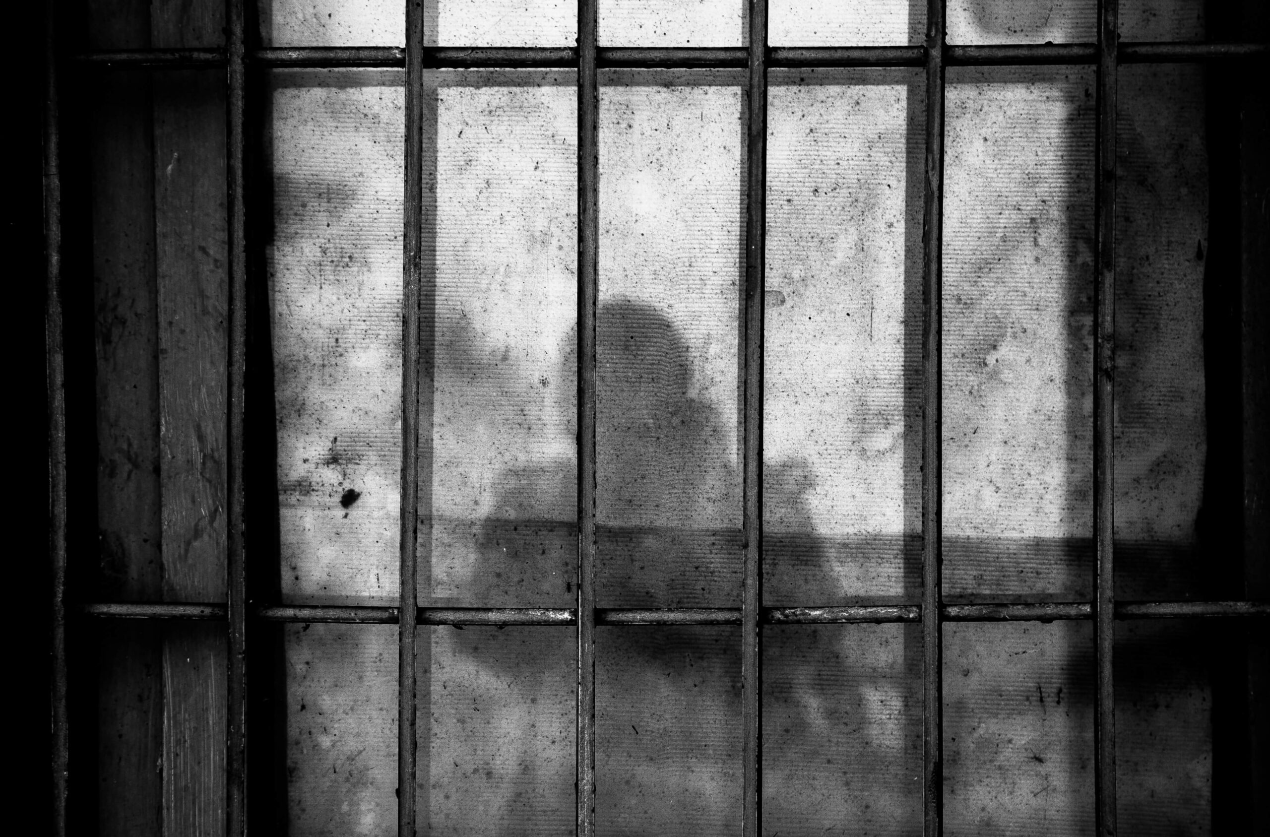 A black and white image of prison bars against a plain wall and the shadow of a person