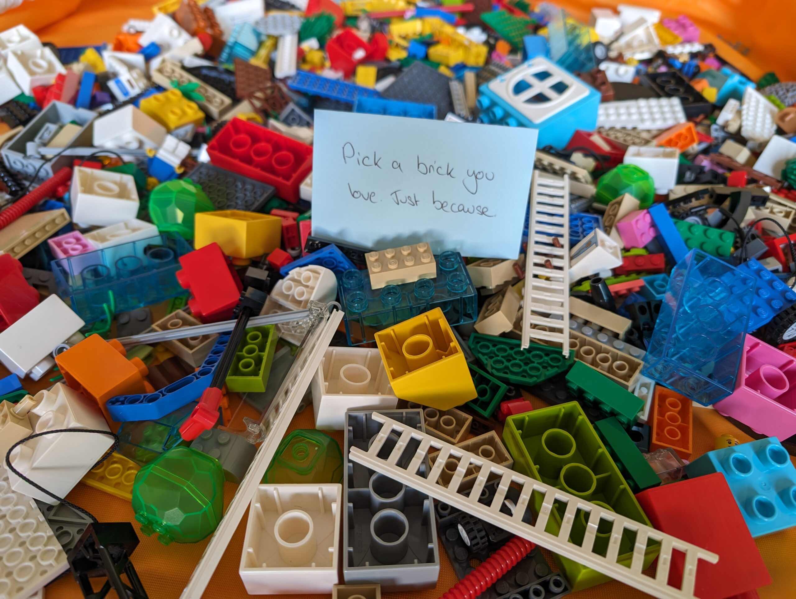 A pile of colourful mixed LEGO bricks with a card on the top that reads "pick a brick you like, just because"