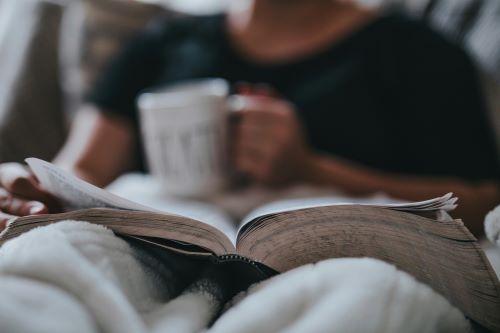 A close up photo of someone balancing a mug on a book while snuggled under a blanket